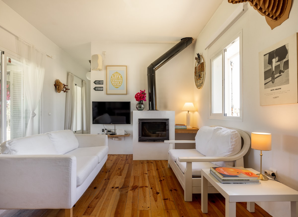 A bright living area features two white sofas placed around a light wooden coffee table. A simple fireplace adds warmth, while a flat-screen TV is mounted on the wall. Natural light flows in through the windows, highlighting the wooden flooring.