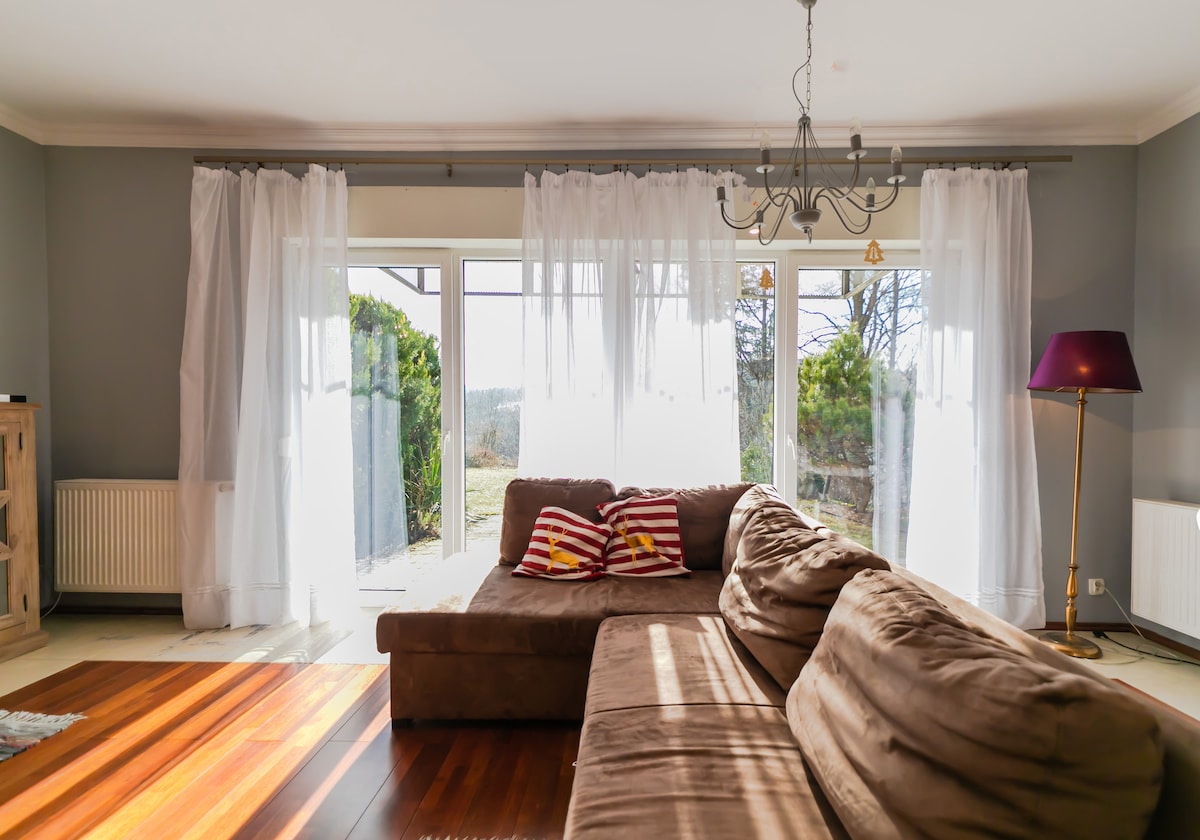 A comfortable living area features a spacious brown sofa with striped cushions. Large glass doors allow natural light to enter, offering views of the outdoors. Sheer white curtains frame the doors, and a modern lamp stands in the corner, enhancing the serene atmosphere.