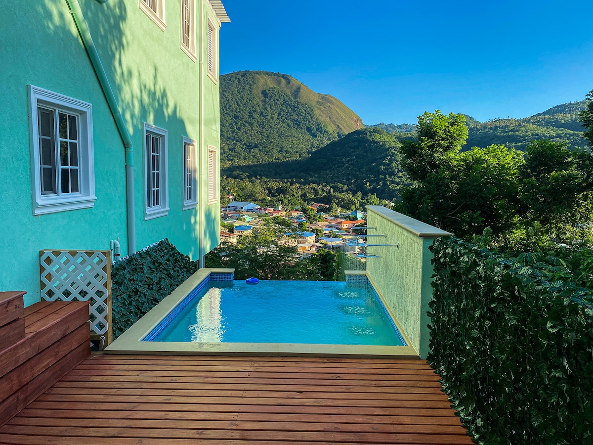 A serene private plunge pool is surrounded by natural wood decking, offering a seamless view of the lush mountains and vibrant town below. The building's light green exterior complements the landscape, while the glass railing provides unobstructed sight lines to the scenic surroundings.