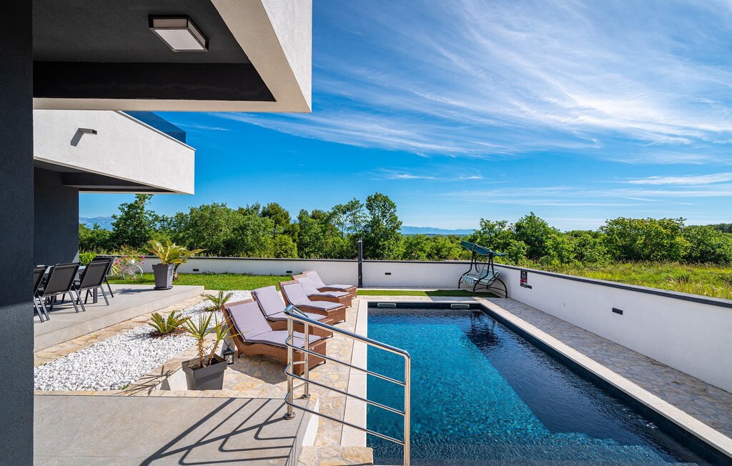An outdoor space is highlighted by a modern swimming pool, flanked by sun-loungers and surrounded by landscaped greenery. The area features a paved patio with a small garden in front of the pool, and a seating area positioned for scenic views of the landscape.