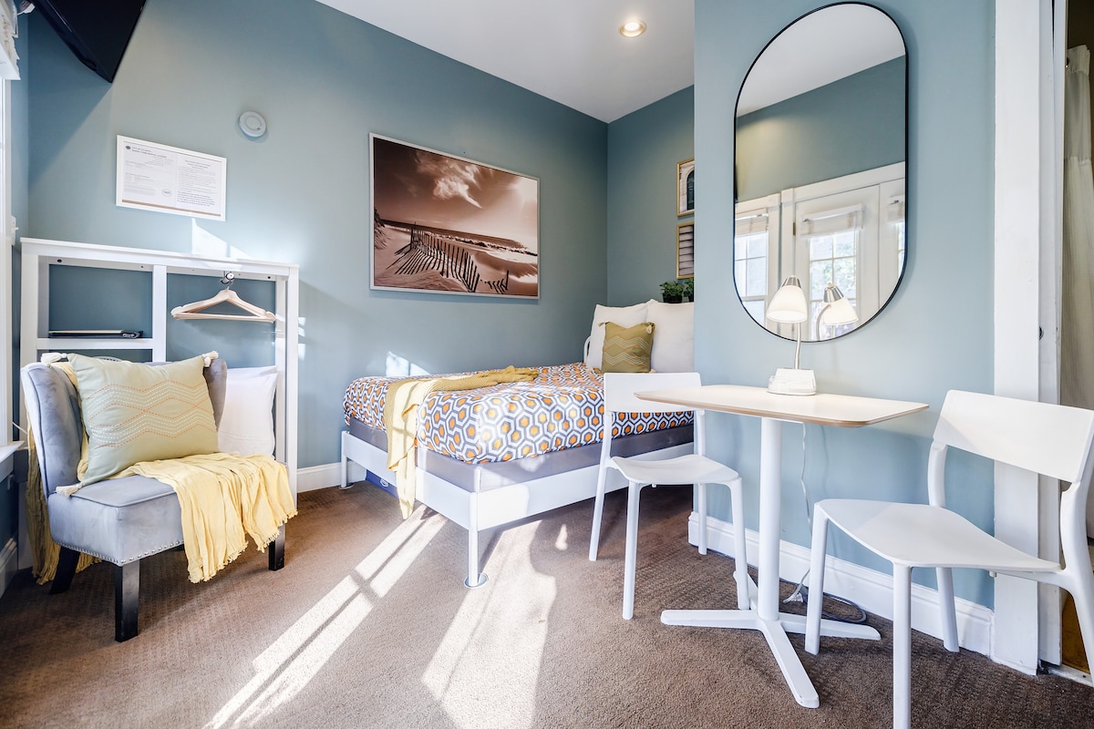 A small studio space features a comfy queen bed with a patterned duvet and a single light blanket. A round mirror hangs above a small dining table paired with two white chairs, while a cozy chair and a clothes rack are positioned nearby. Light enters through a window, illuminating the room.