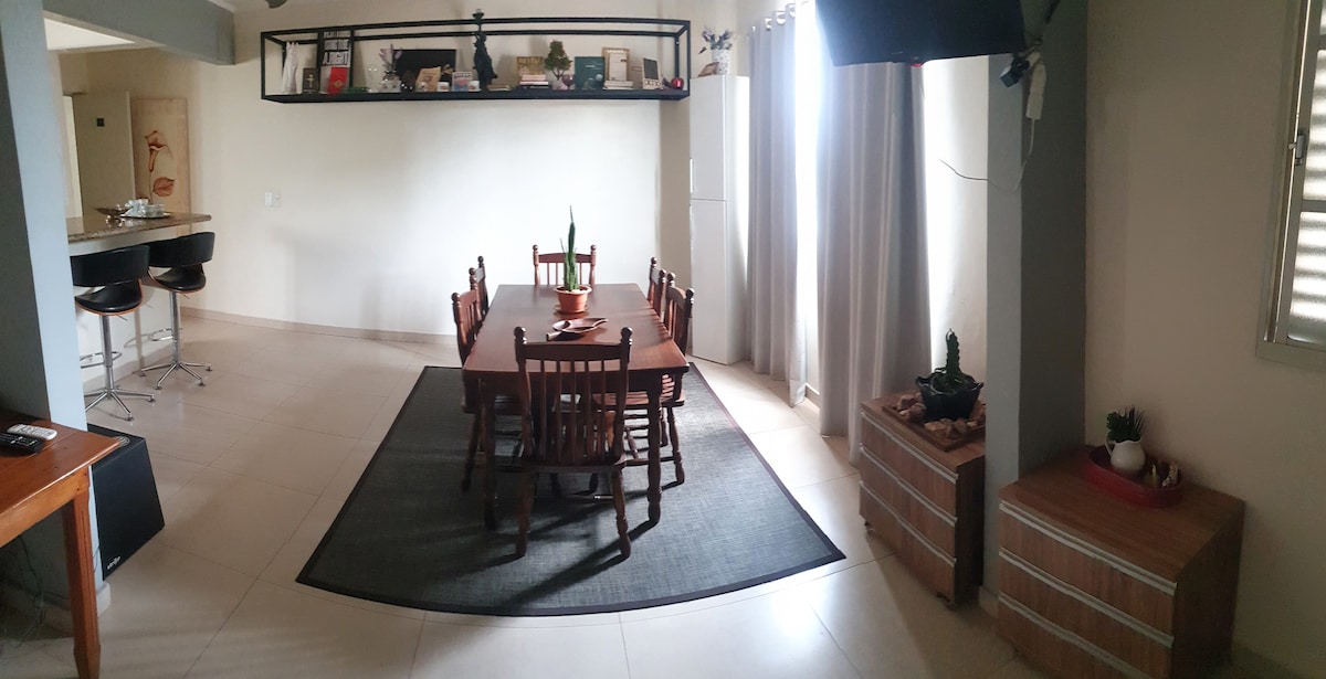 A welcoming dining area is featured with a large wooden table surrounded by several chairs. A dark area rug anchors the space, while natural light filters through sheer curtains. Shelves above the table hold decorative items, adding a touch of personality to the room.