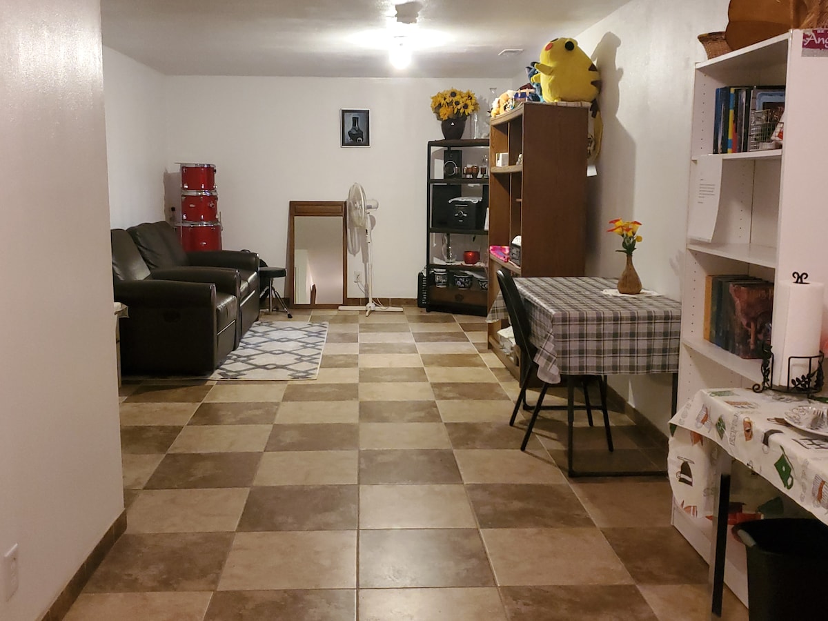 A spacious living area features a sectional sofa facing a wall-mounted television. A table with a checkered tablecloth is paired with a chair. Decorative items include a flower vase and a bookshelf filled with books. The flooring consists of patterned tile in a checkered design.
