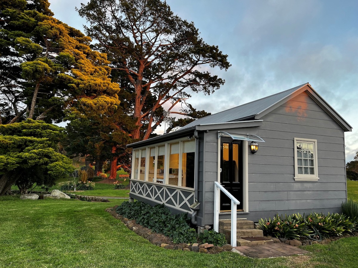 The studio cottage showcases a charming exterior with light gray siding and a metal roof. Surrounded by well-maintained gardens, it features large windows allowing natural light, complemented by a welcoming entryway. Tall trees and lush greenery enhance the scenic country setting.