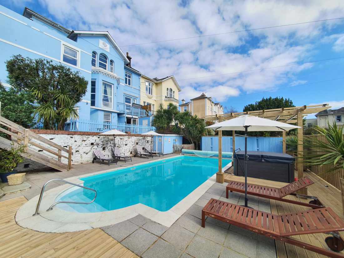 A serene swimming pool is prominently featured in the outdoor area, surrounded by sun loungers and parasols. The guest house, painted in light blue, rises in the background amid lush greenery. The sky is partly cloudy, adding a tranquil ambiance to the scene.