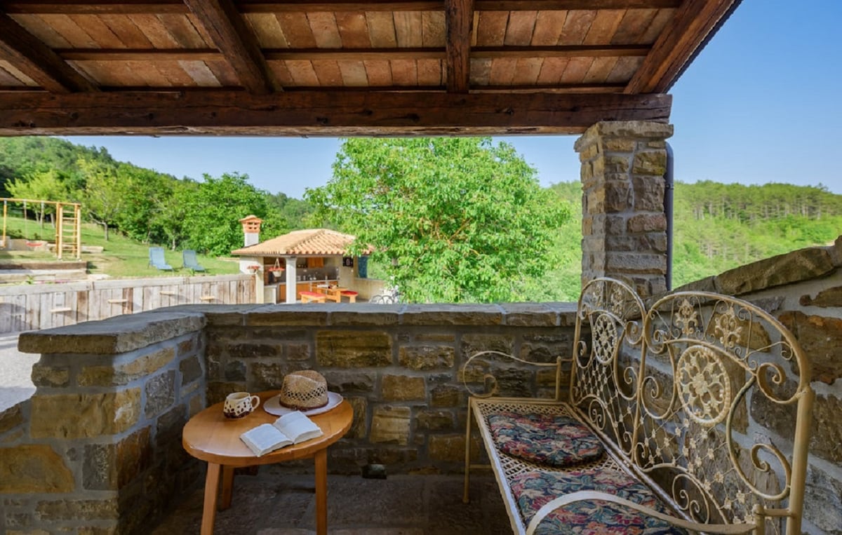 A shaded seating area features a decorative wrought-iron bench and a small wooden table. The view includes lush greenery and a stone structure, with sunlight filtering through the outdoor space, enhancing the natural elements surrounding the area.
