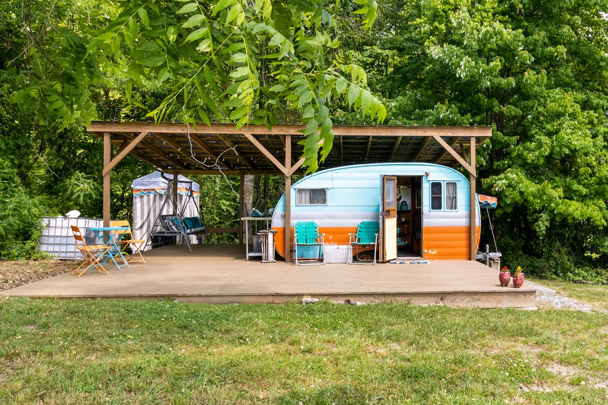 A vintage canned ham camper is positioned under a spacious covered deck surrounded by lush green foliage. Outdoor seating includes several teal chairs and a foldable table, providing a relaxed space. The camper entrance is open, revealing a glimpse of the inviting interior.