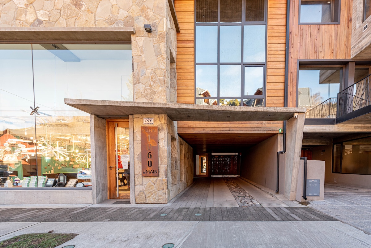 The entrance of the building is characterized by a blend of stone and wood, highlighted by large glass windows. A covered passage leads to the interior, showcasing a modern architectural design. The area is well-lit, enhancing the welcoming atmosphere.