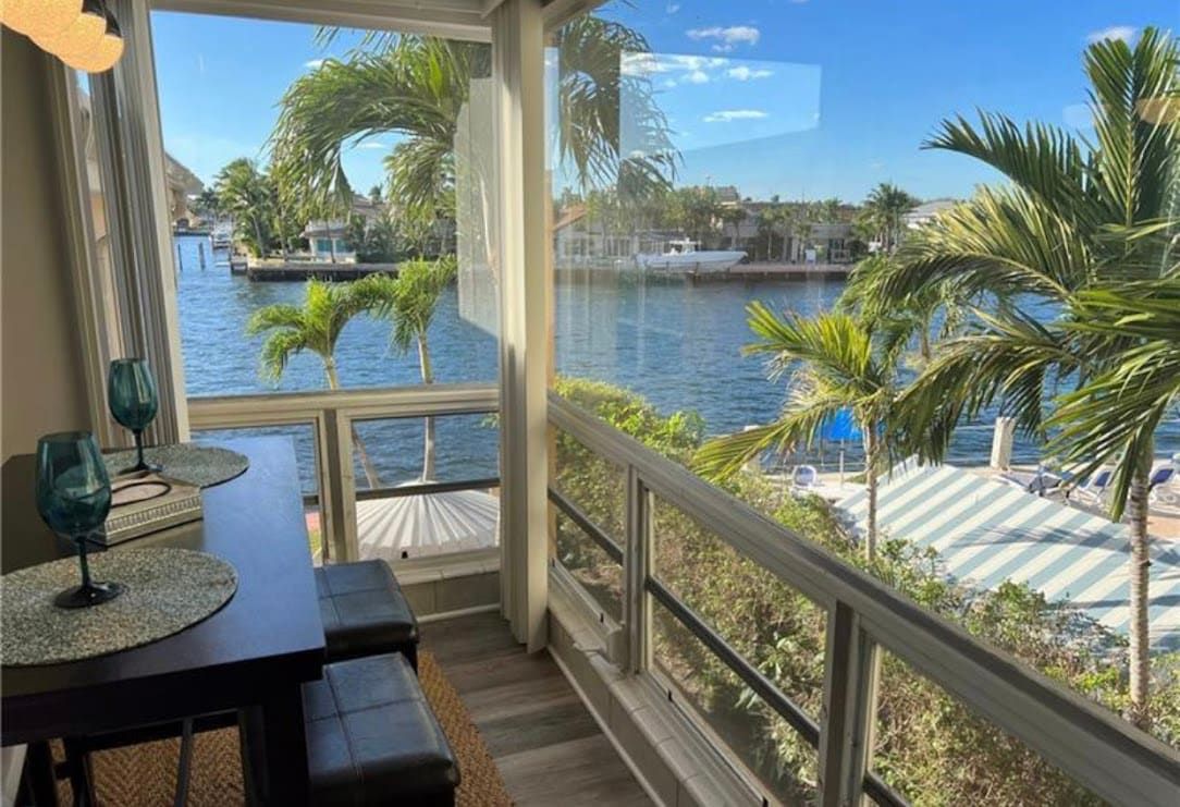 A dining nook is positioned by large windows, offering a view of the intercoastal waterway lined with palm trees. A dark wood table is accompanied by two sleek chairs, inviting relaxation while enjoying the scenic surroundings.