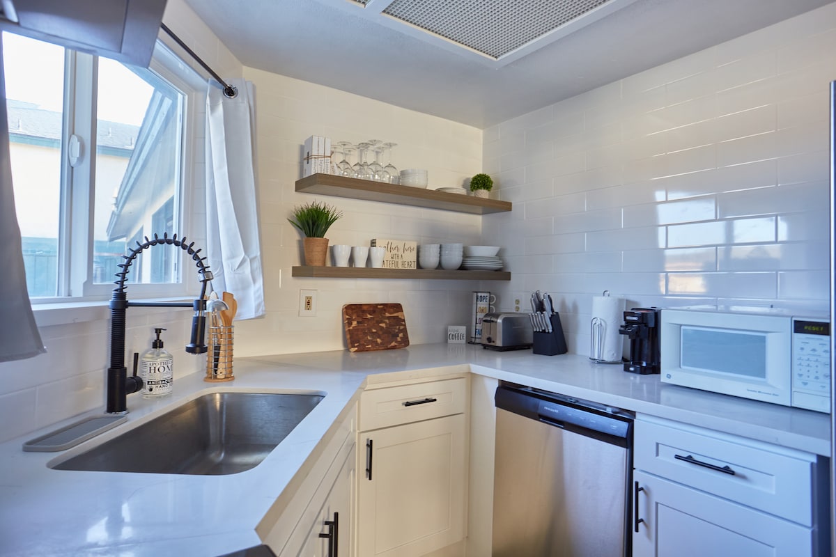 The kitchen features modern white cabinetry and sleek countertops. A stainless steel sink is paired with a stylish faucet. Open shelving displays glassware and decorative items, while essential appliances, including a microwave and coffee maker, are positioned for convenience.