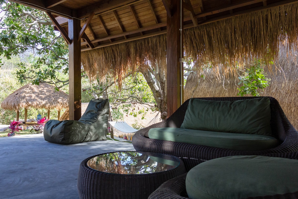 A shaded outdoor seating area features comfortable, oversized bean bag chairs and a low glass table, surrounded by lush greenery. A gently thatched roof adds to the serene ambiance, while a hammock can be seen nearby, inviting relaxation in a peaceful setting.