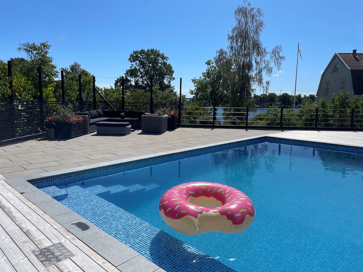 The image showcases a pool area surrounded by a deck, featuring a pink donut-shaped float resting on the clear water. Lush greenery and trees line the background, while lounge seating is visible nearby, creating a relaxing outdoor space.