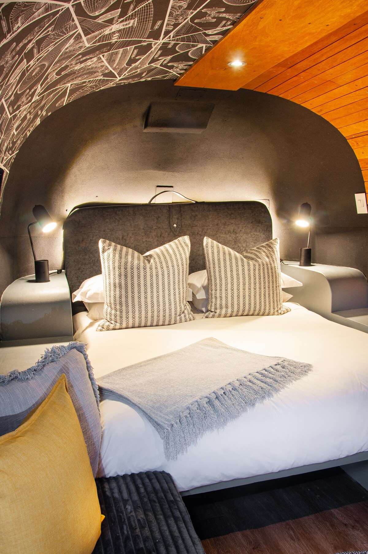 A cozy sleeping area features a comfortable bed with decorative pillows and a soft throw blanket. Ambient lighting is provided by two bedside lamps. The arched walls are adorned with unique artwork, creating an intimate atmosphere. A tasteful yellow accent pillow adds a pop of color.
