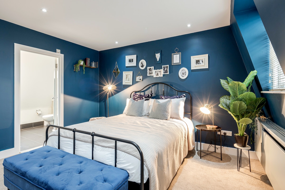 A spacious bedroom features a large bed dressed in neutral linens, complemented by decorative pillows. A cozy sitting area is provided by a tufted ottoman, while a floor lamp and potted plant add warmth. Gallery-style wall art enhances the room's aesthetic.