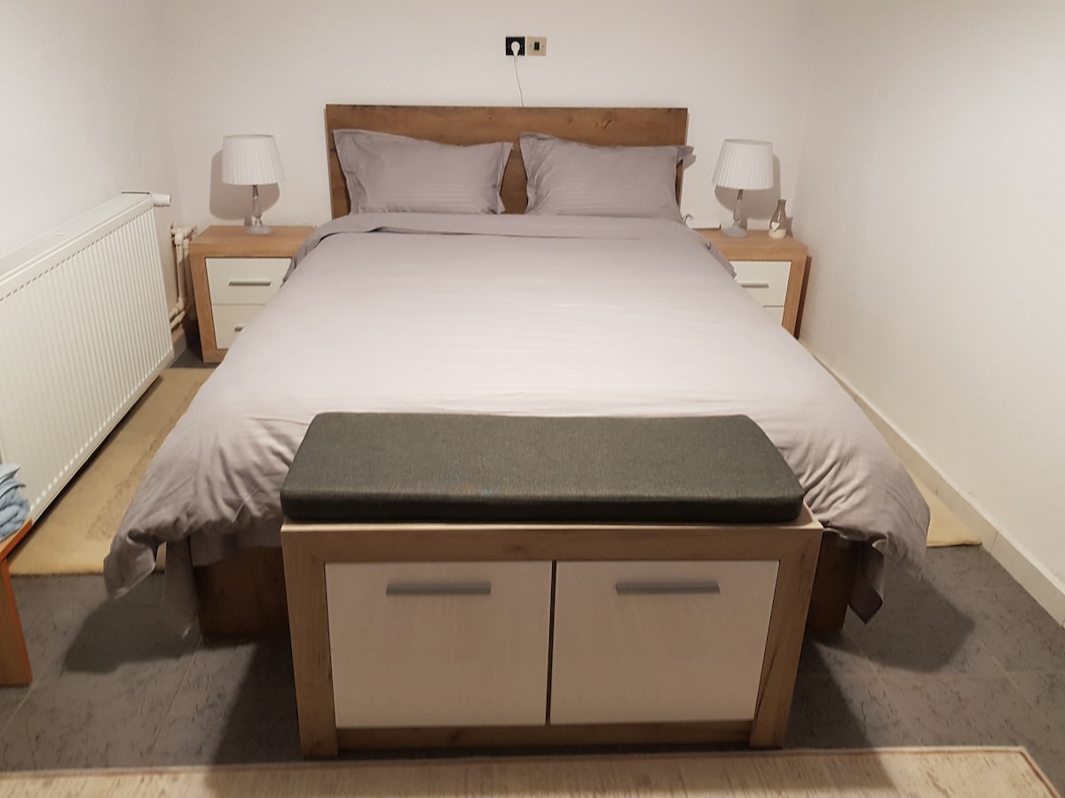 A cozy bedroom features a spacious bed with neutral-colored bedding, flanked by two bedside lamps. Light wooden furniture, including nightstands and a bench at the foot of the bed, complements the calming atmosphere. A radiator is positioned under the window for added comfort.