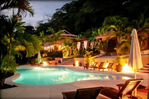 A serene outdoor pool area is surrounded by lush tropical foliage and illuminated by soft lighting in the evening. Comfortable lounge chairs are positioned around the water, while wooden steps lead to bungalows nestled amid the greenery.