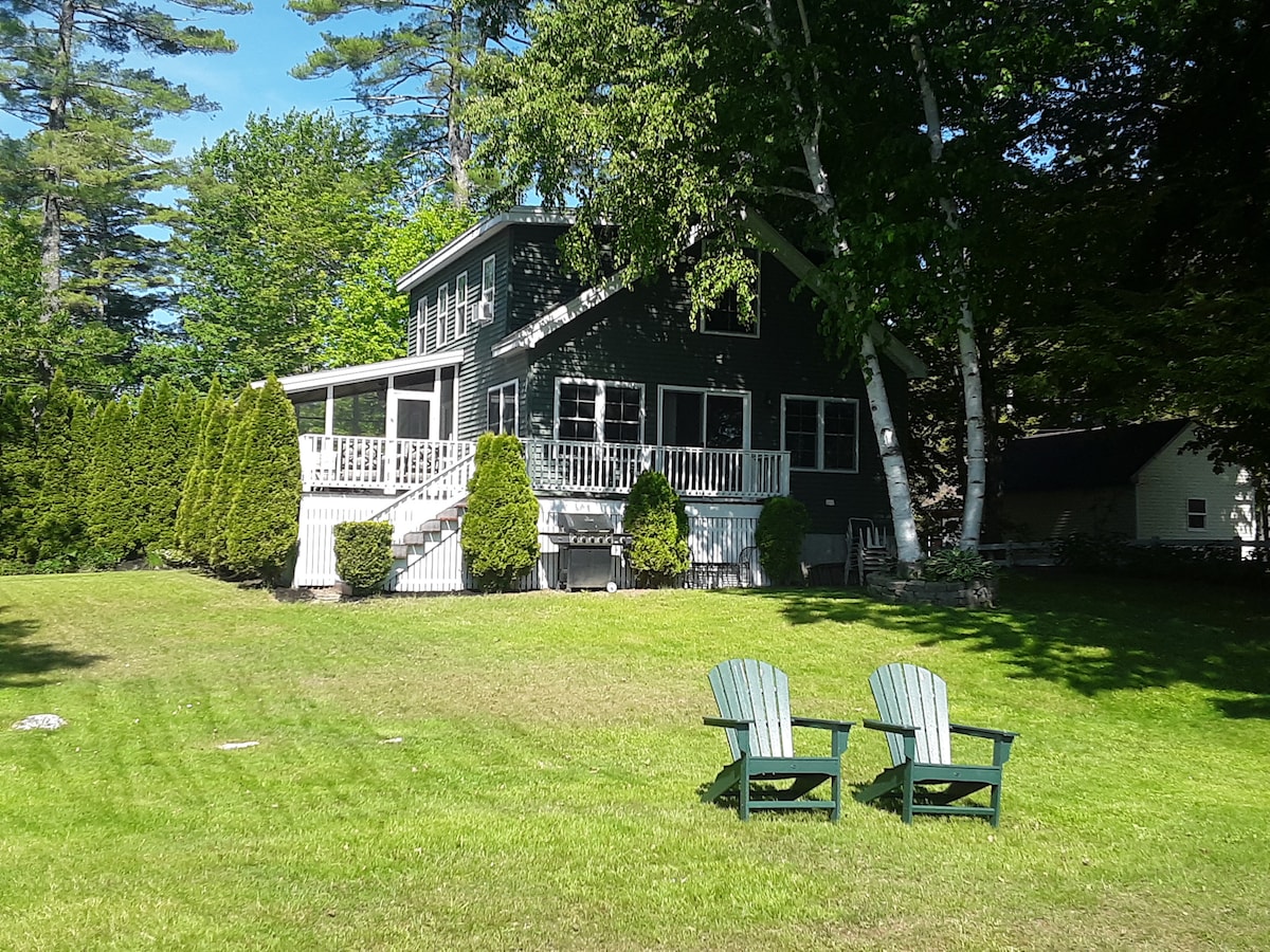 A well-maintained two-story home is surrounded by lush greenery and tall trees. A spacious lawn is present in front, featuring two Adirondack chairs facing the house. A large deck with white railings is visible on the upper level, providing outdoor seating options.