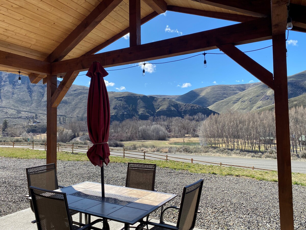 A covered porch features a dining table surrounded by several chairs. Above, string lights are softly arranged, illuminating the outdoor space. In the background, rolling hills and dry vegetation create a scenic view under a clear blue sky.