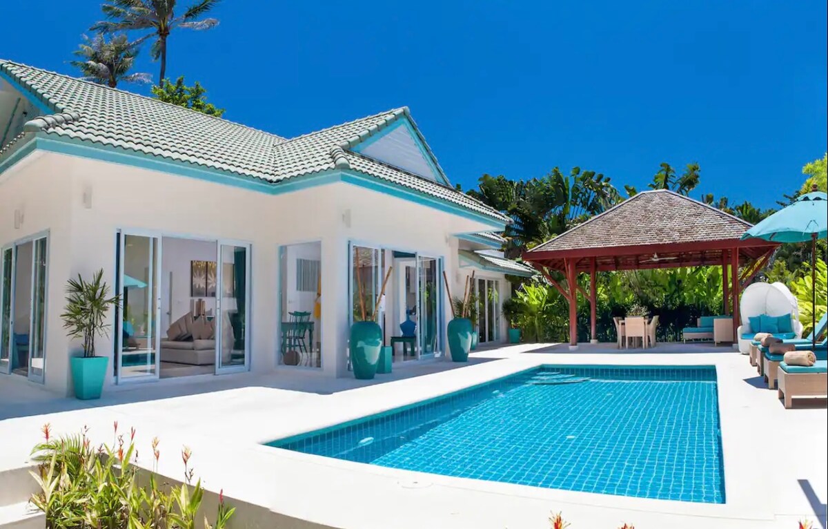 A spacious villa is showcased with a private swimming pool at the center. Surrounding the pool are sunbeds and a covered dining area, complemented by vibrant turquoise accents. Lush greenery frames the villa, enhancing the tranquil outdoor setting under clear blue skies.