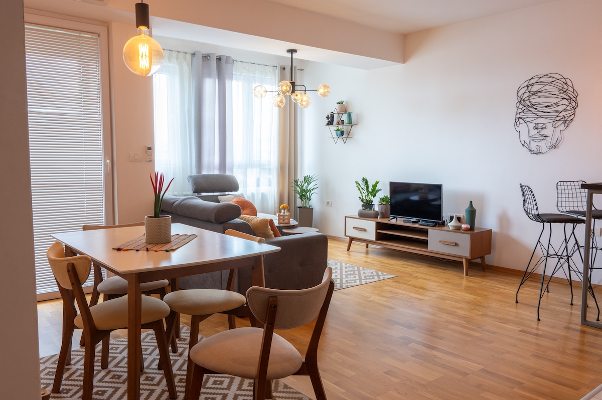 An inviting living space features a dining table with wooden chairs, a comfortable sofa, and a flat-screen TV. Natural light pours in through large windows, illuminating decorative plants placed around the room. A stylish wall art piece adds a contemporary touch to the neutral decor.