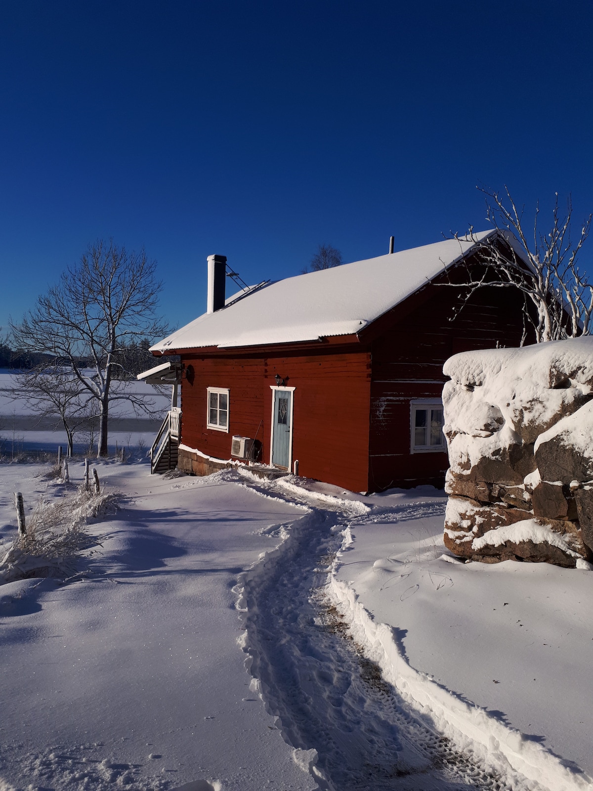 A red wooden cabin is surrounded by a blanket of snow, with a clear blue sky overhead. A visible pathway, marked by footprints, leads to the entrance, while a stone wall provides contrast against the fresh snowfall.