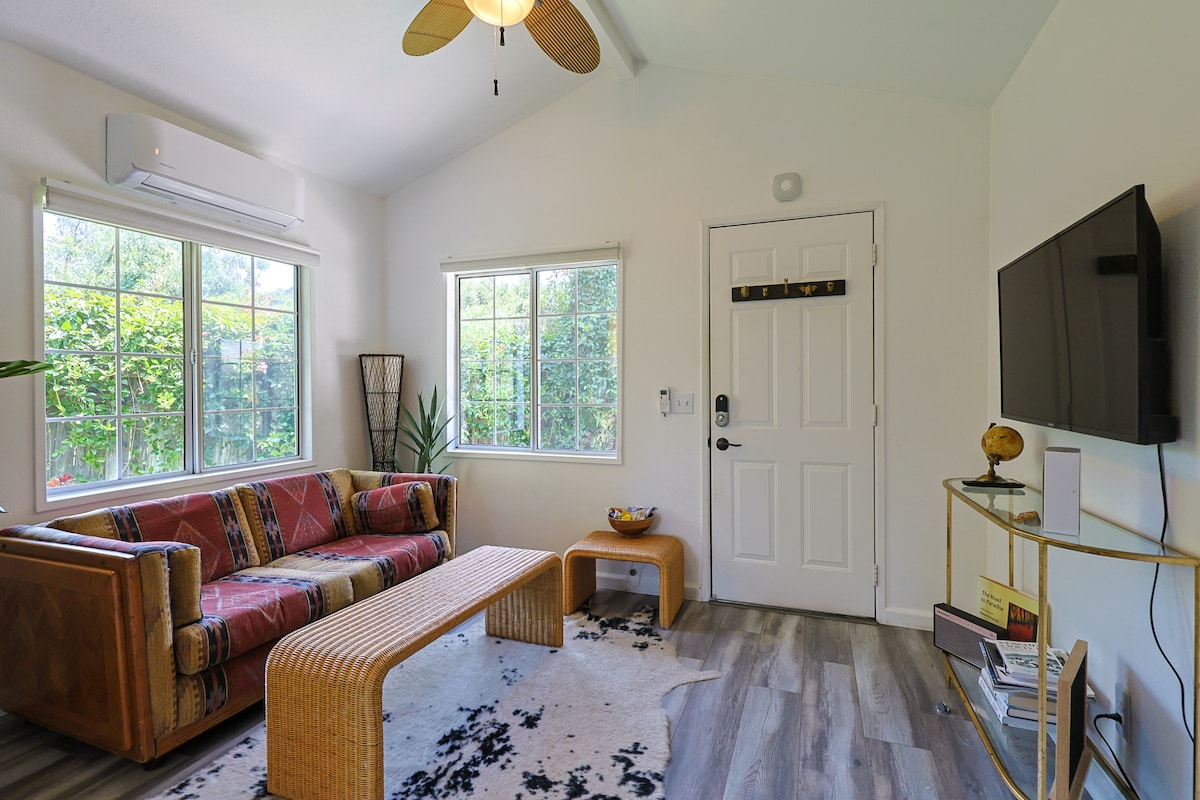 A bright and airy living area features large windows allowing natural light to fill the space. A comfortable seating arrangement includes a woven coffee table and a textured area rug. The room is complemented by a wall-mounted TV and tropical plants, adding a touch of nature.