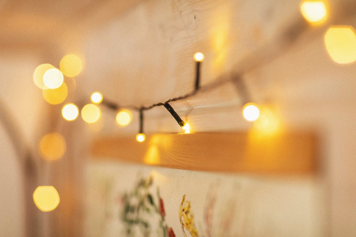 Soft, warm lighting from string lights is draped above a decorative wall hanging. The lights create a gentle glow, enhancing the cozy ambiance of the interior space.
