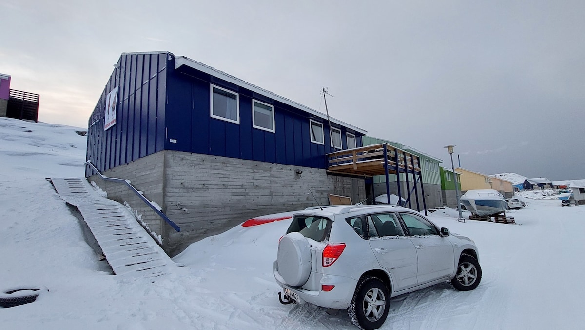 A modern blue structure with multiple windows is set against a snowy landscape, showcasing a sloped ramp leading to the entrance. A parked white vehicle is positioned in front, providing a sense of accessibility to the building.