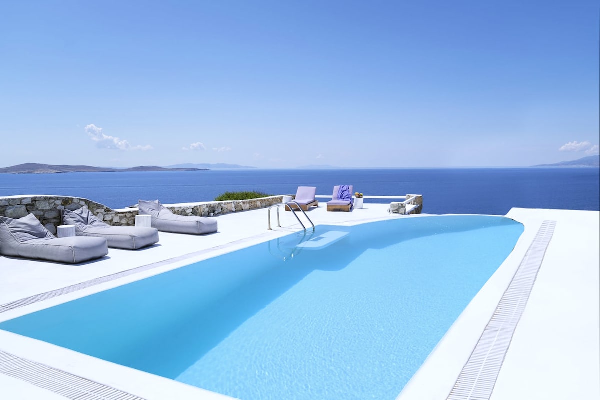 An infinity-edged swimming pool stretches across the terrace, reflecting the clear blue sky. Comfortable sunbeds line the pool area, offering views of the Aegean Sea. The surrounding stone walls frame the space, enhancing the sense of tranquility and connection with nature.
