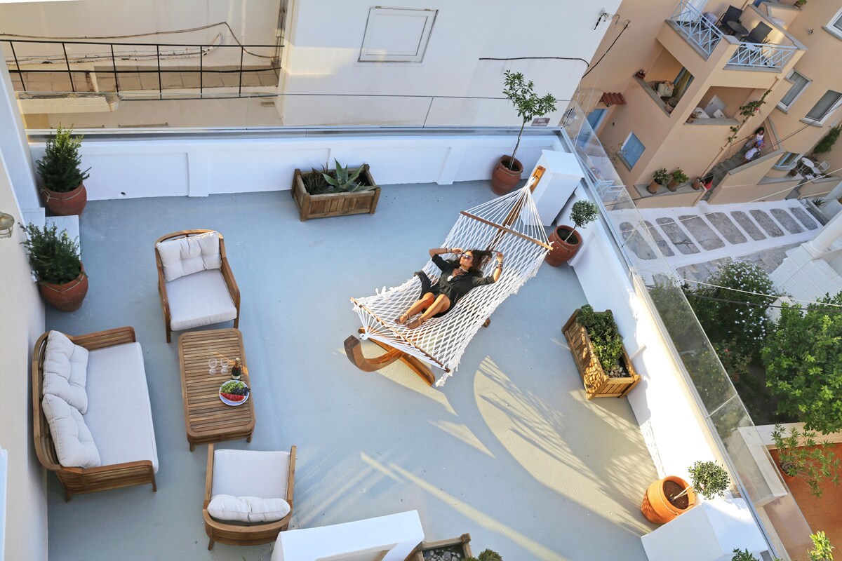 A spacious private terrace features comfortable seating options, including a lounge area with wicker chairs and a hammock. Potted plants are arranged along the perimeter, enhancing the serene outdoor space, which offers a view of the surrounding buildings and alleyways.