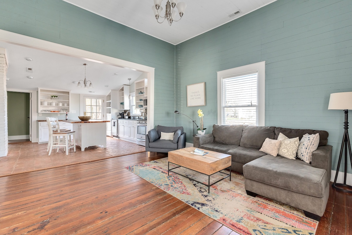 A spacious living room features a large lounge-style couch alongside a coffee table, all set on original hardwood floors. Natural light filters through the windows, highlighting the soft green walls. An open layout connects to a dining area and kitchen, contributing to a welcoming atmosphere.