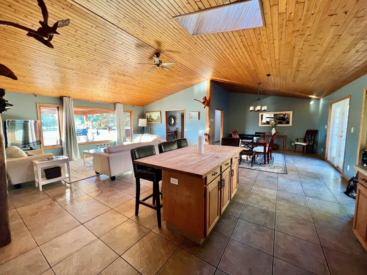 A spacious interior is highlighted by a wooden ceiling with skylights. An open layout includes a kitchen island, living area, and dining space. Large windows offer natural light and views of the outdoor scenery. Comfortable seating arrangements and warm-toned decor create an inviting environment.