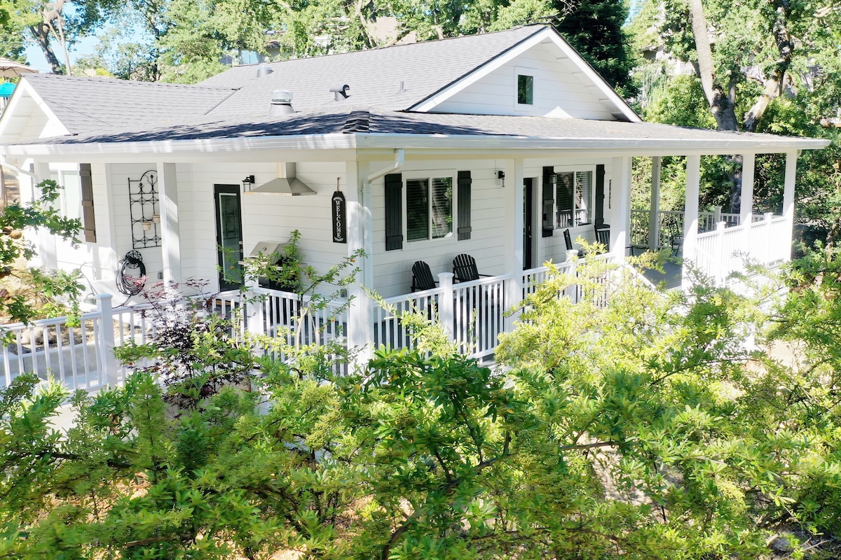 A welcoming exterior view of a single-story guesthouse is displayed, showcasing a wrap-around covered porch. The porch features rocking chairs, a grill, and lanterns, surrounded by lush greenery and trees that provide a serene, natural setting.