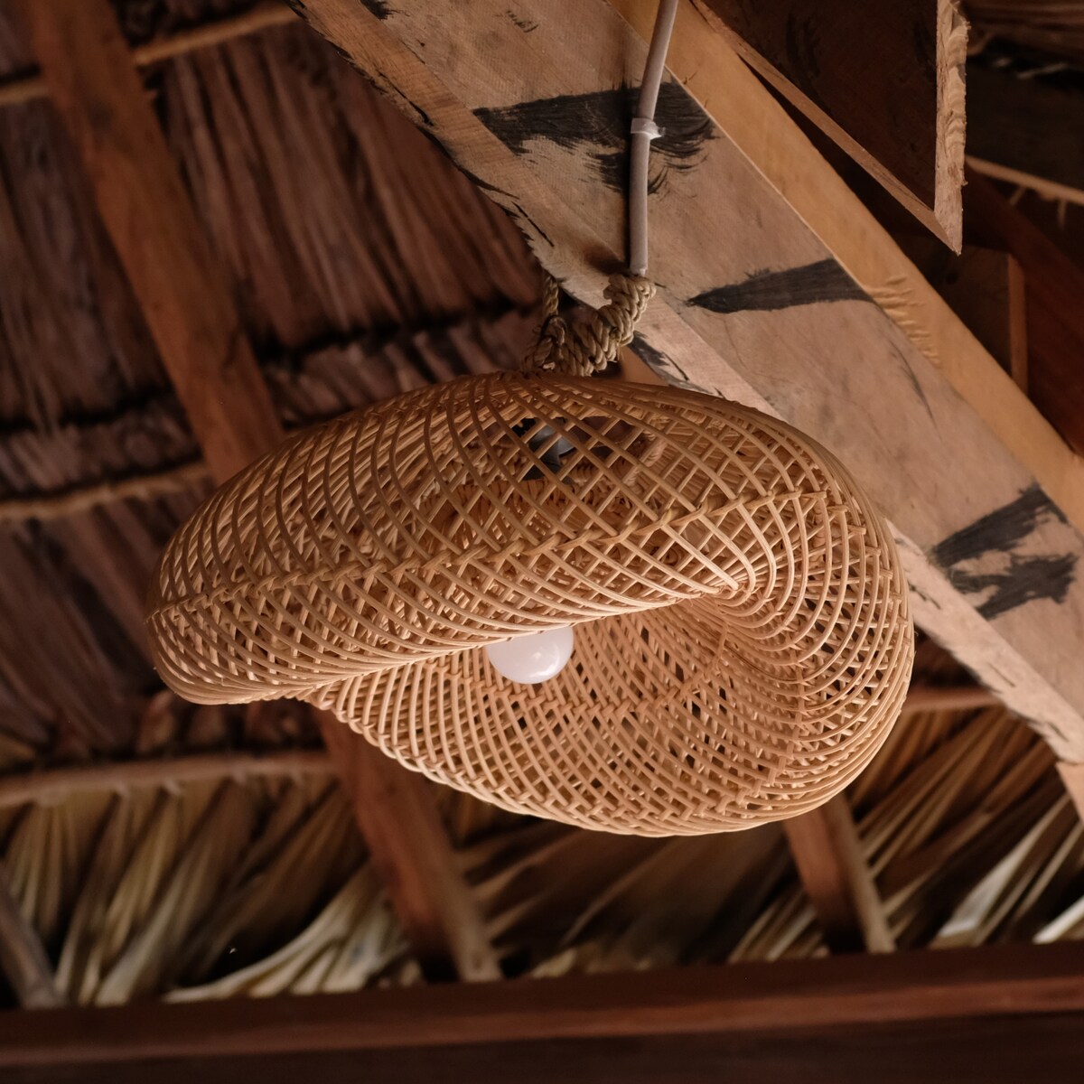 A woven pendant light hangs from the wooden ceiling beams, showcasing an intricate design made of natural materials. The soft light bulb is visible within the basket-like structure, blending organic aesthetics with functionality, emphasizing the earthy atmosphere of the space.
