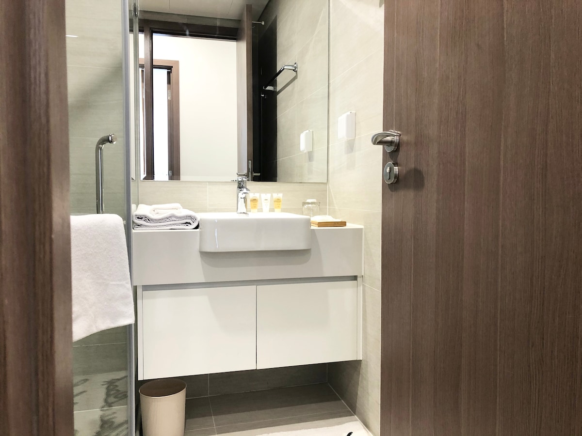 A modern bathroom is equipped with a white vanity and a square sink. The space features a mirrored cabinet above the sink, glass shower enclosure, and neutral tiled walls. Towels are neatly arranged on the counter, with personal care products placed nearby.