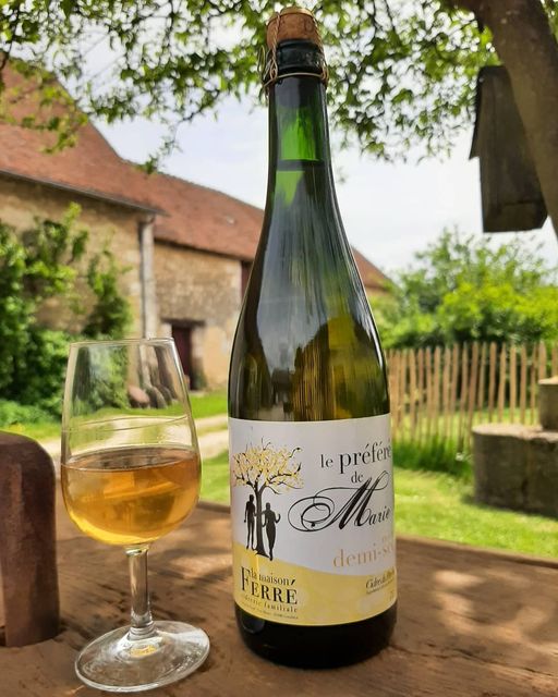 A glass of golden sparkling cider is positioned on a wooden table, reflecting sunlight. The bottle displays a label featuring a tree design and the name 'le préféré de Marie.' A rustic farmhouse and vibrant greenery are visible in the background.