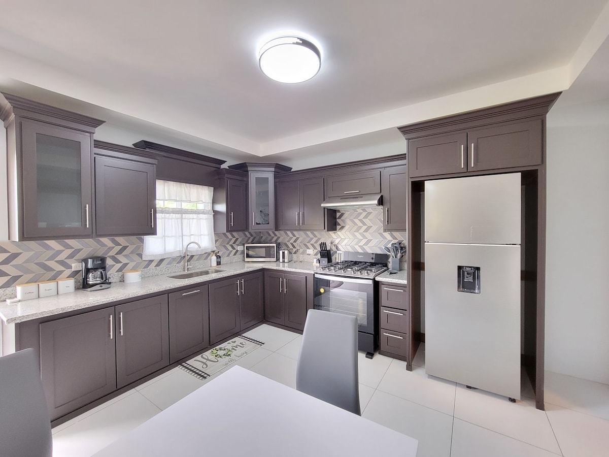 The kitchen features modern cabinetry in a rich brown hue, complemented by a stainless steel refrigerator and gas stove. Light-colored countertops are paired with patterned backsplash tiles, while ample natural light brightens the space through a nearby window.