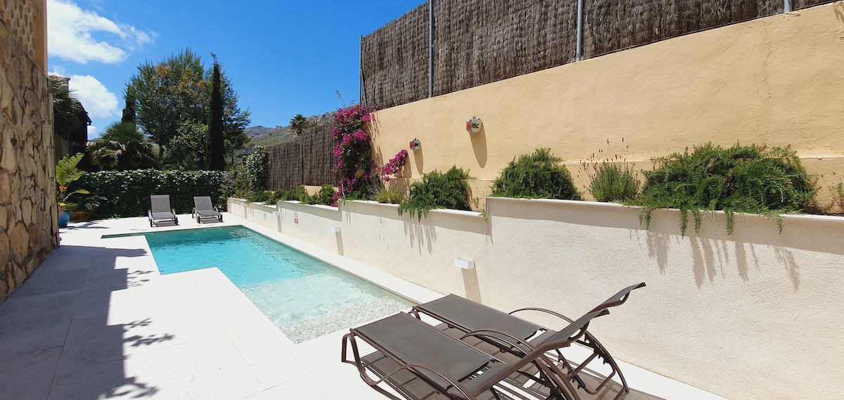 A serene outdoor space features a private swimming pool surrounded by smooth white tiles. Two sun loungers are positioned beside the pool, offering a place for relaxation. Vibrant flowers climb the wall, adding a touch of color against the beige backdrop under a bright blue sky.