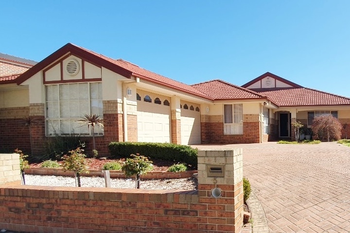 The exterior of a spacious two-story house is displayed, featuring a classic brick facade and a red-tiled roof. A paved driveway leads to the garage, and meticulously arranged garden beds with shrubs and decorative stones enhance the curb appeal.