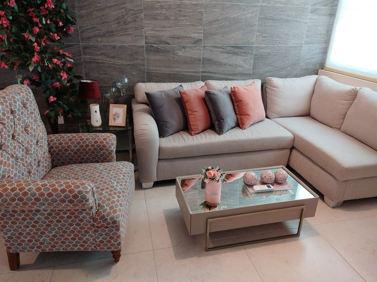 A comfortable living area features a light-colored sectional sofa adorned with coordinating pillows. A patterned armchair complements the seating arrangement. A glass coffee table, accented by decorative items including flowers, is positioned in the center, enhancing the inviting atmosphere of the space.