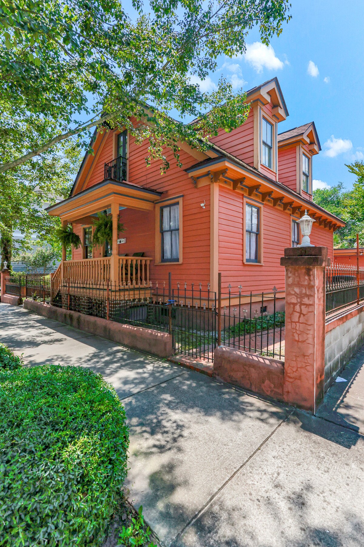 A charming orange house with a classic architecture features a welcoming front porch surrounded by lush greenery. A wrought iron fence encloses the property, and paved walkways lead to the entrance, enhancing the home's street appeal.