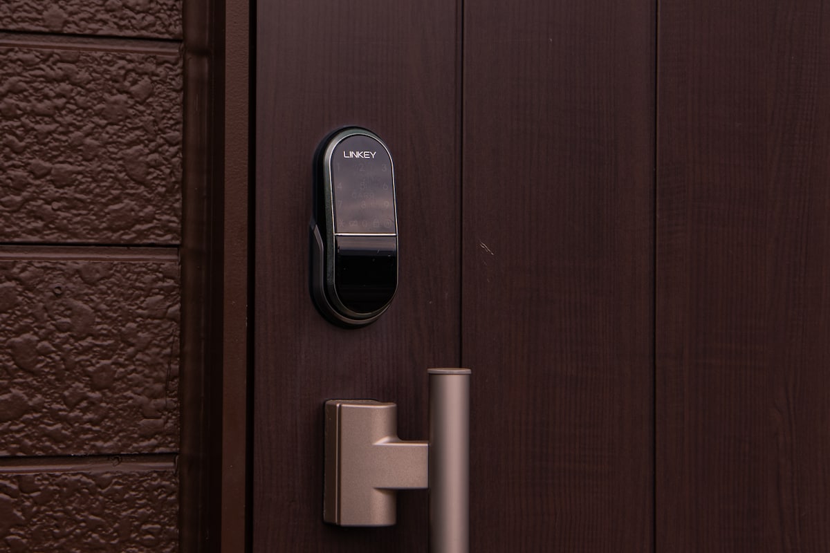 The entrance door is adorned with a sleek, modern electronic lock, designed for keyless entry. A polished silver handle complements the dark, textured wood paneling of the door, creating an inviting entrance.