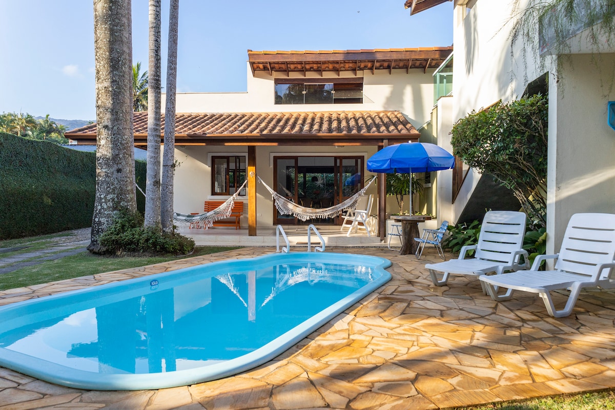The inviting pool area features a sparkling blue pool surrounded by a tiled deck. Two white lounge chairs are positioned near the water, complemented by an umbrella and hammocks strung between palm trees, creating a relaxing outdoor environment.
