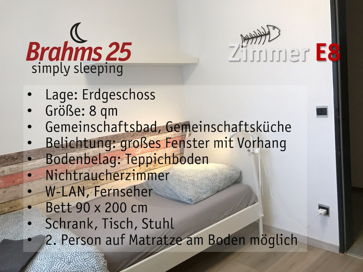 A simply furnished room features a spacious bed with a light-colored blanket. A large window with curtains allows natural light. The floor is carpeted, and basic furniture includes a wardrobe, table, and chair. The room is set up for non-smokers and includes a television.