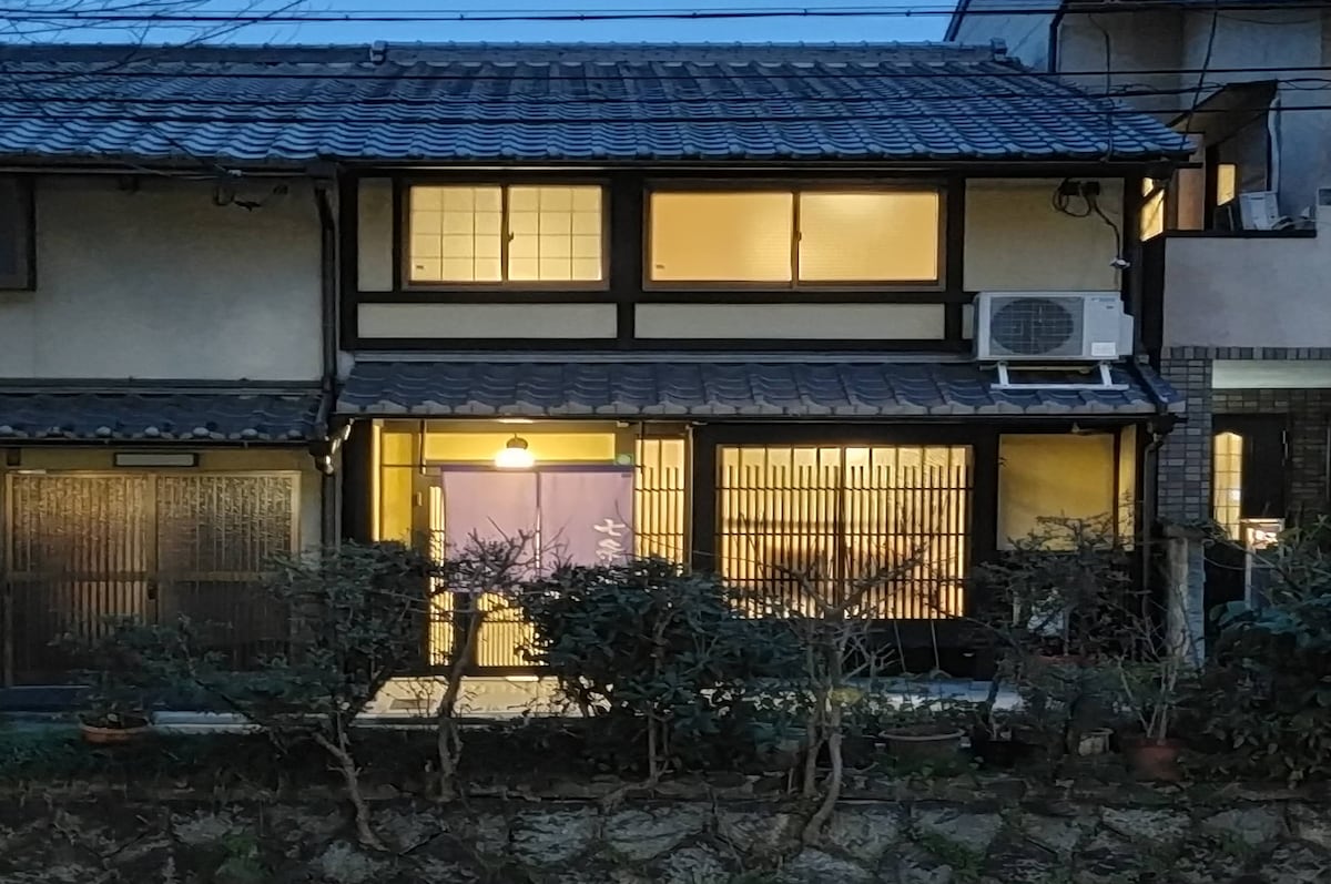 A traditional two-story Japanese townhouse is illuminated at dusk, revealing a warm atmosphere through large glass doors. The exterior features wooden beams and a tiled roof, complemented by greenery in the foreground. Air conditioning units are visible on the side wall.