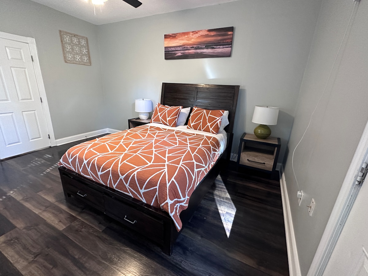 A cozy bedroom is equipped with a queen bed featuring an orange and white geometric-patterned duvet. Two bedside tables with lamps are positioned on either side. The floor showcases dark hardwood, and light enters through a window, accentuating a calming atmosphere.