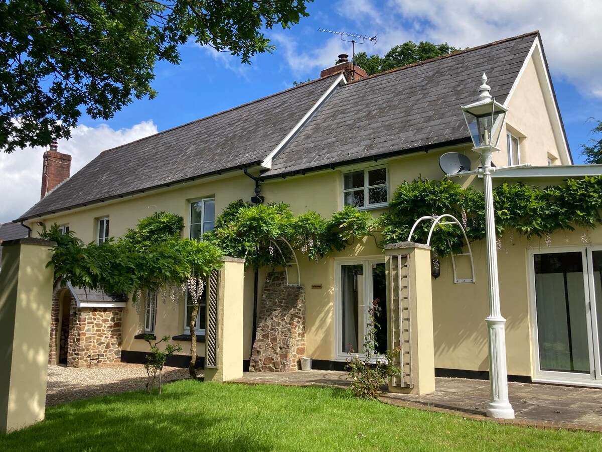 A spacious farmhouse exterior is visible, featuring a light yellow facade complemented by stone accents. Lush greenery frames the entrance, and large glass doors provide access to the interior. A serene garden area is present in the foreground, offering a peaceful setting.