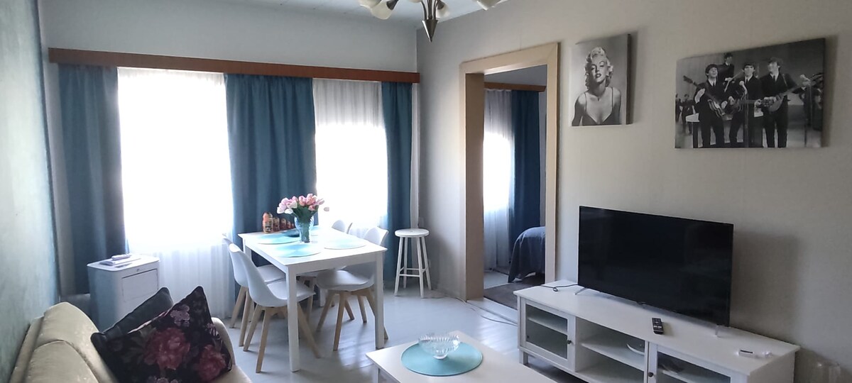 A bright living area features large windows with light blue curtains, allowing natural light to illuminate the space. A white dining table is set for four, accompanied by a cozy couch with decorative pillows. A television rests on a low media unit, enhancing the inviting ambiance.