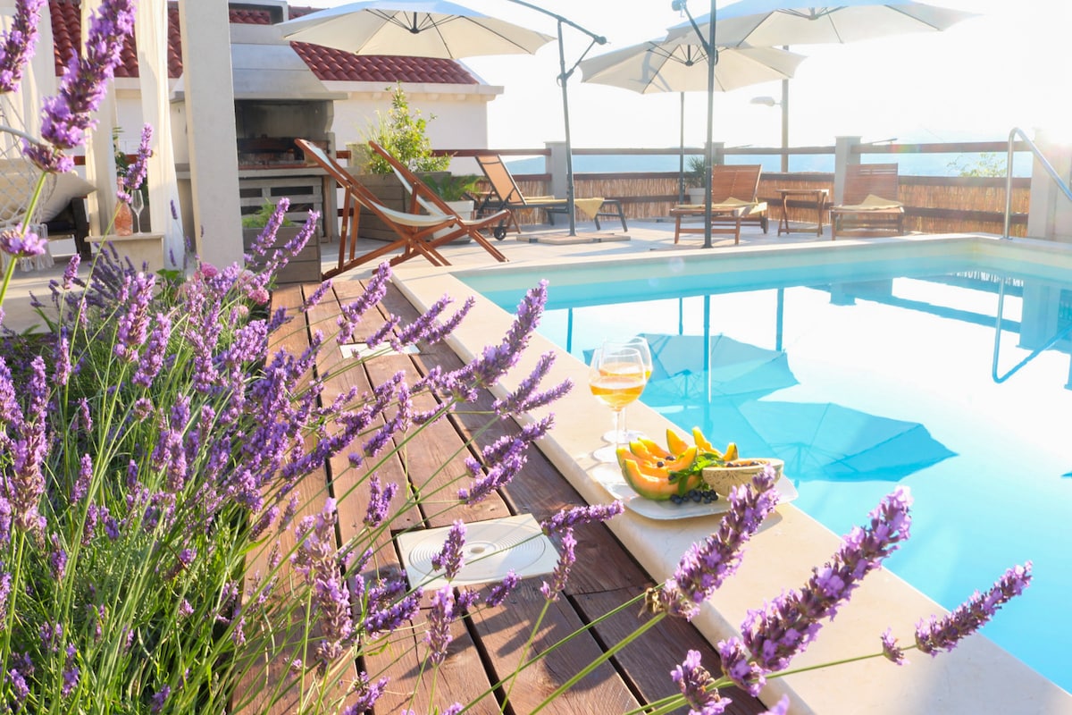 A serene outdoor space is highlighted by a swimming pool reflecting the sun. Lavender flowers border the wooden deck, while umbrellas provide shade over sun loungers. A coffee table holds a refreshing drink and fruit, set against a backdrop of the sea and distant hills.
