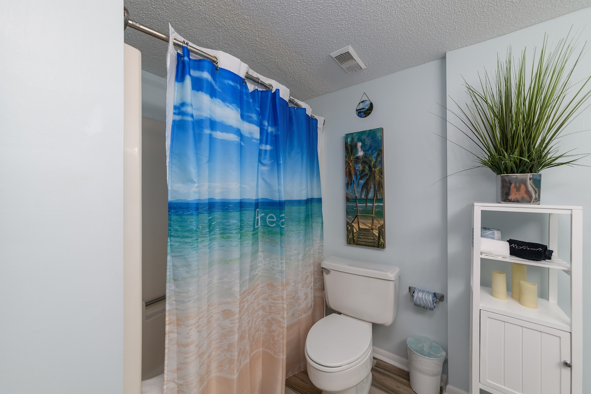 A bathroom is shown, featuring a light blue wall color and a beach-themed shower curtain depicting serene ocean waves. A toilet is positioned next to a small white shelf adorned with towels and decor. A decorative plant adds a touch of greenery.